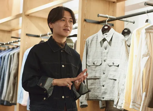 トップスが並ぶ前で、接客しているタカハラさん